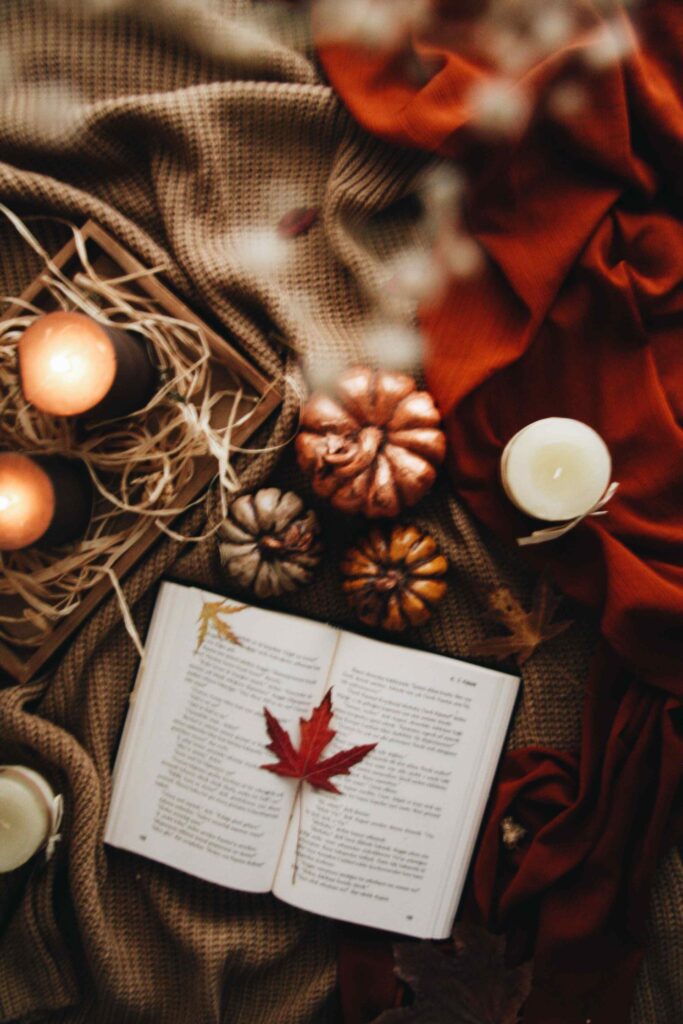 Cozy autumn reading nook featuring an open book, lit candles, decorative pumpkins, soft knit blankets, and warm fall accents for a relaxing seasonal atmosphere.